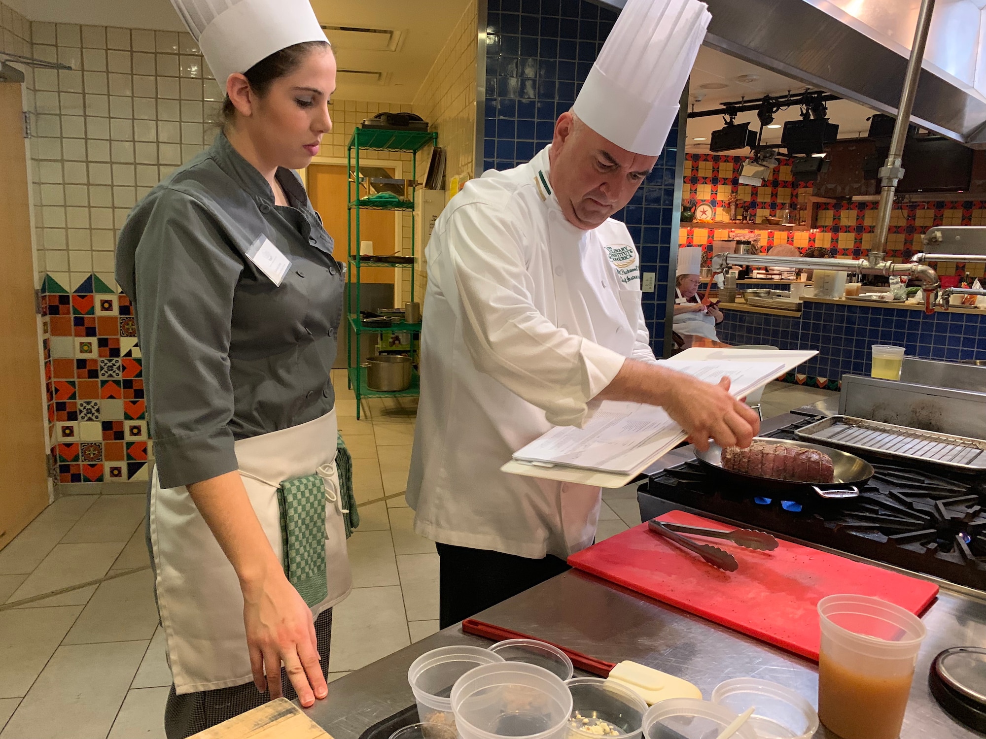 Culinary Institute of America San Antonio Chef Will Packwood and Gabriela Irwin, a chef at Spangdahlem Air Base, Germany, look over a pork loin recipe during the practical portion of the first Air Force Techniques of Healthy Cooking Course June 5, 2019, in San Antonio. Sixteen nonappropriated fund food and beverage chefs and cooks from across the Air Force attended the course established by the Air Force Services Center, in partnership with the CIA San Antonio. The five-day course is designed to teach and demonstrate healthier ingredients, flavorful spicing and better cooking methods to deliver healthier menu choices at food service operations across installations, not just at dining facilities. (U.S. Air Force photo by Debbie Aragon)