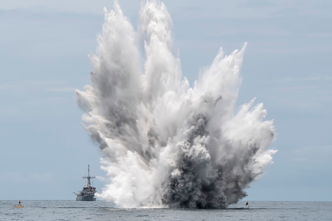 A controlled mine detonation in the ocean sends water high into the sky as a Navy ship observes from a distance.