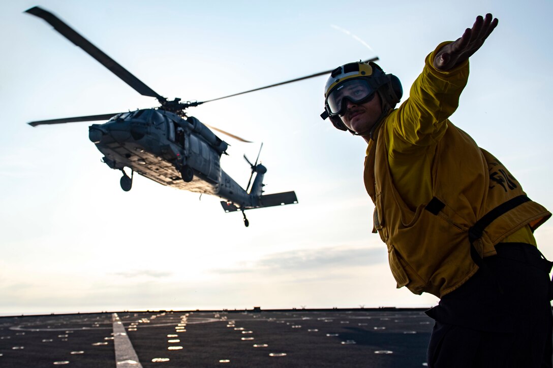 A sailor wearing a yellow vest, helmet and goggles signals as a helicopter hovers over a ship’s flight deck.