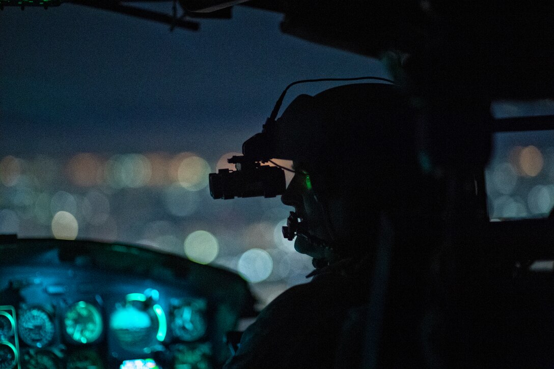 An airman sits inside of a plane wearing a helmet and goggles at night.