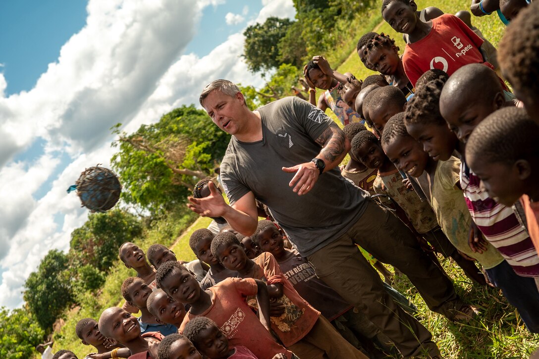 A soldier in civilian clothes throws a ball in the air as children gathered around him watch.