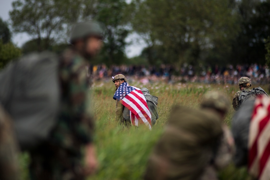 A U.S. paratrooper walks with a U.S. flag on his shoulders after conducting a static-line jump into Iron Mike Drop Zone outside of Saint-Mere-Eglise, France June 9, 2019. Paratroopers conducted the largest airdrop since Operation Neptune June 6, 1944. (U.S. Air Force photo by Senior Airman Devin M. Rumbaugh)