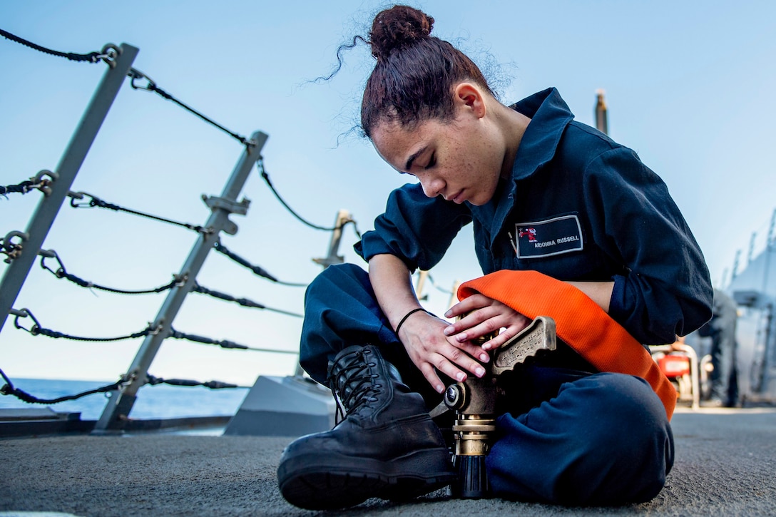 A sailor connects a fire hose while sitting cross-legged on the deck of a ship.