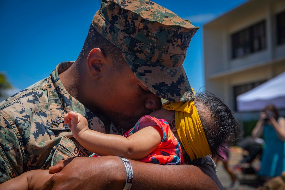 A sailor in a camouflage uniform and cover kisses and holds a baby in his arms.