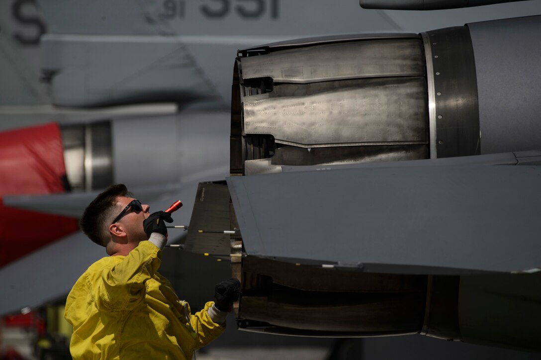 An airman wearing a yellow jacket and sunglasses points a flashlight at a jet engine.