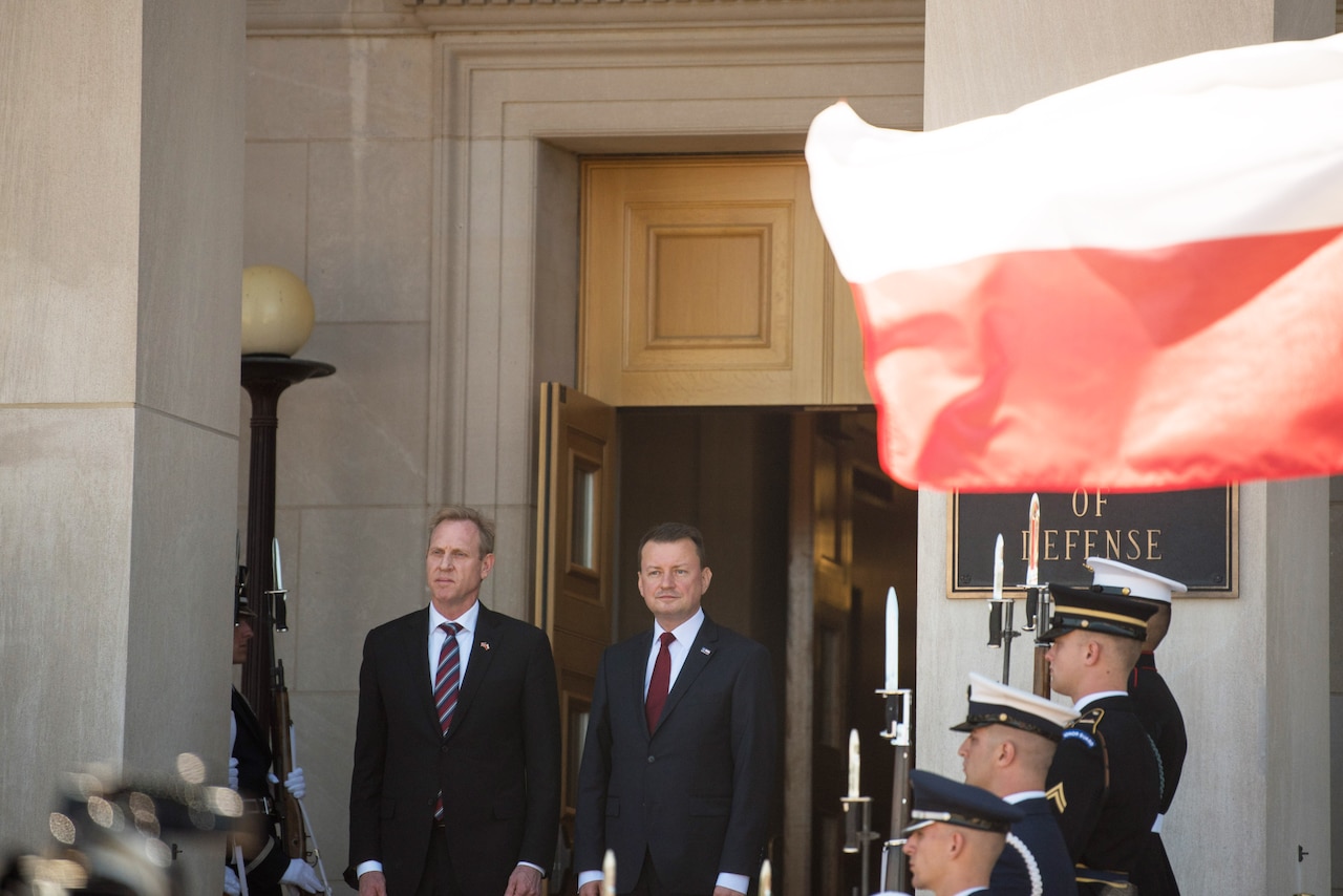 Acting Defense Secretary Patrick M. Shanahan stands with another leader at the top of a staircase flanked by service members as a Polish flag flies.