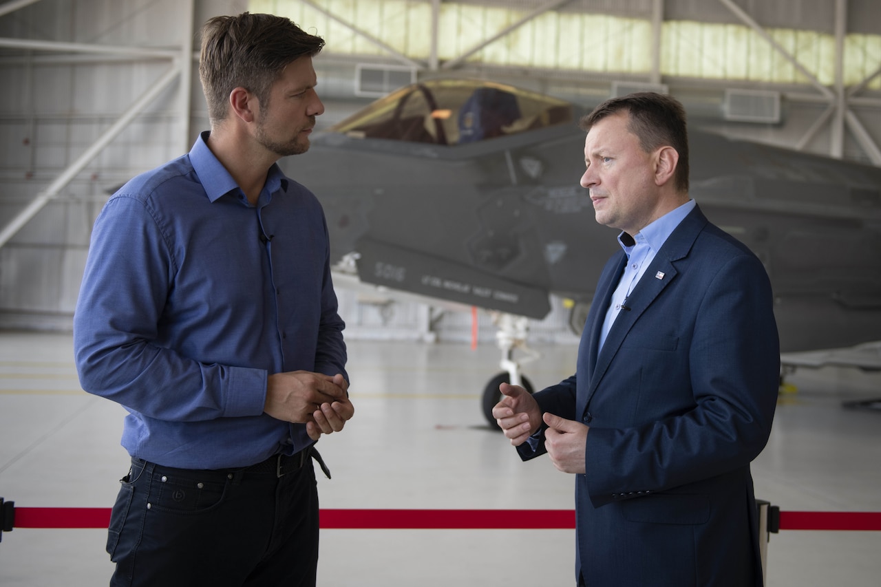 Two men speak inside an aircraft hangar with a fighter aircraft in the background.