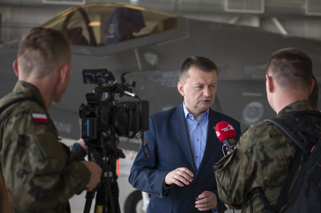 A man is interviewed on camera in an aircraft hangar with a fighter aircraft in the background.