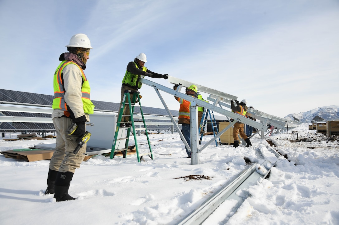 Men wearing winter clothes and hard hats install metal brackets in snowy conditions.