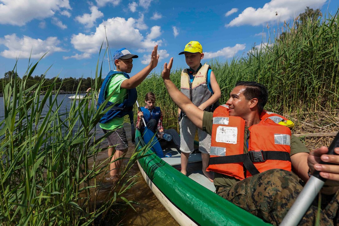 A Marine wearing a life vest in a canoe high-fives a child wading near shore as others look on.
