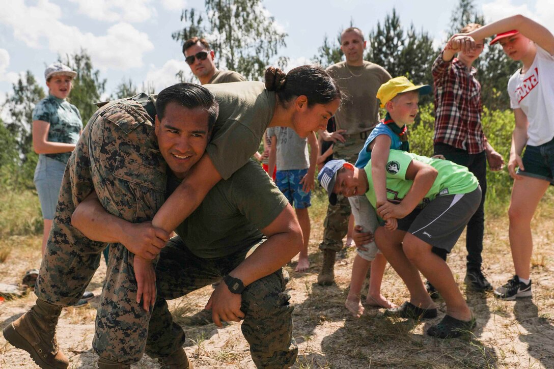 Two Marines demonstrate how to carry a person as children look on and attempt to imitate it.