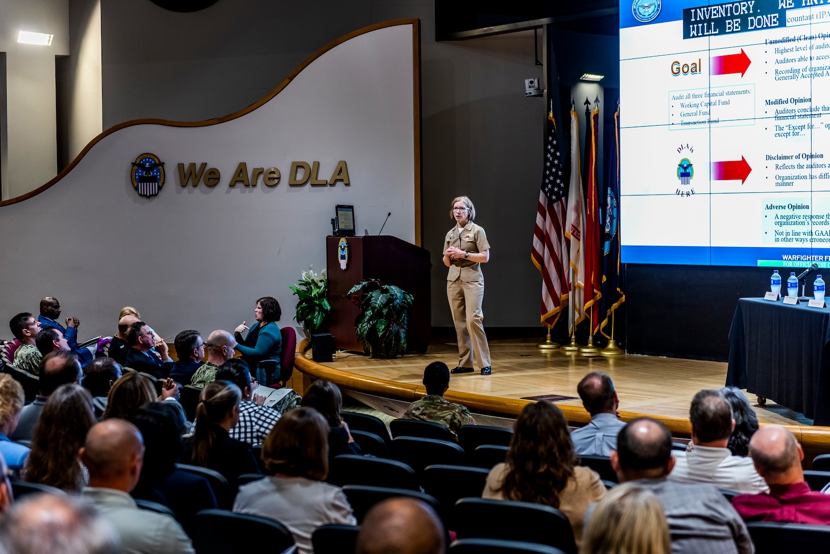 Military woman speaking from a stage to members in the audience.