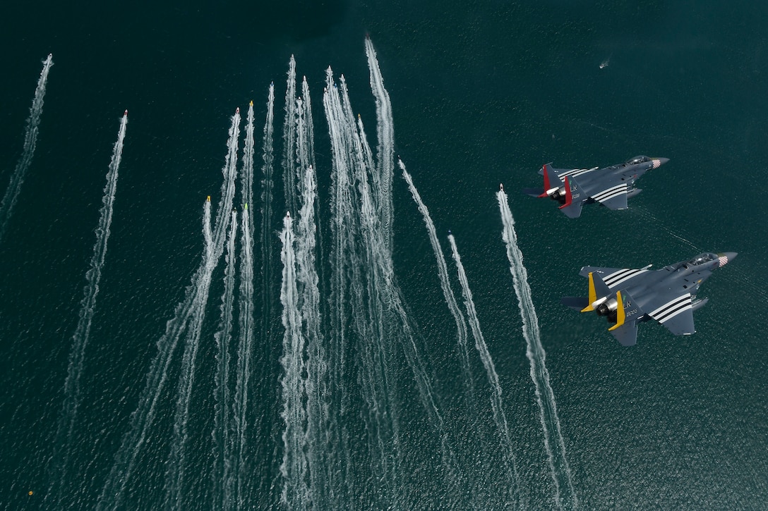 A pair of fighter jets fly above the water where a multitude of boats are moving.