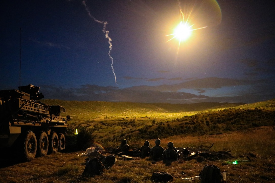 Soldiers sit on the ground at night near an armored vehicle as a sky-borne weapon illuminates a valley.