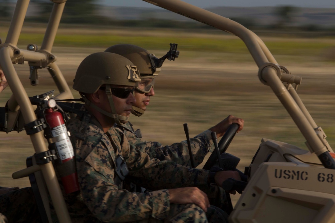 Marines wearing helmets drive an open-air vehicle against a blurry background.