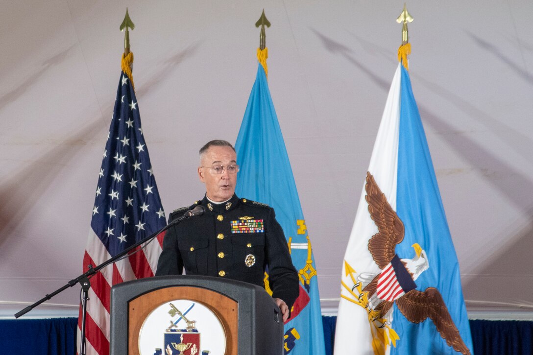 A general speaks at a podium in front of three flags.