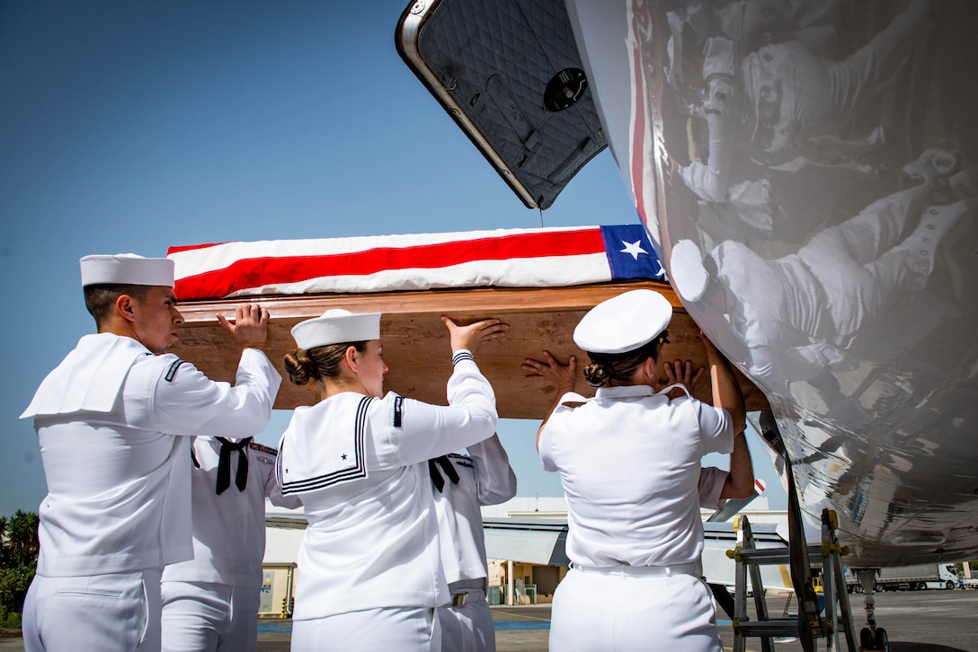 Sailors carry a flag-draped casket into an aircraft.