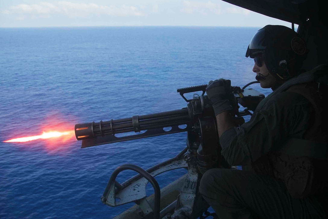 A Marine fires a rotary machine gun from an aircraft flying over the sea.
