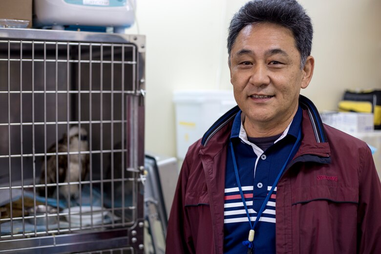 キャンプ・キンザーの環境部に勤務する新崎正紀さんと、動物クリニックで回復中のケージの中のミサゴ（オスプレイ）。