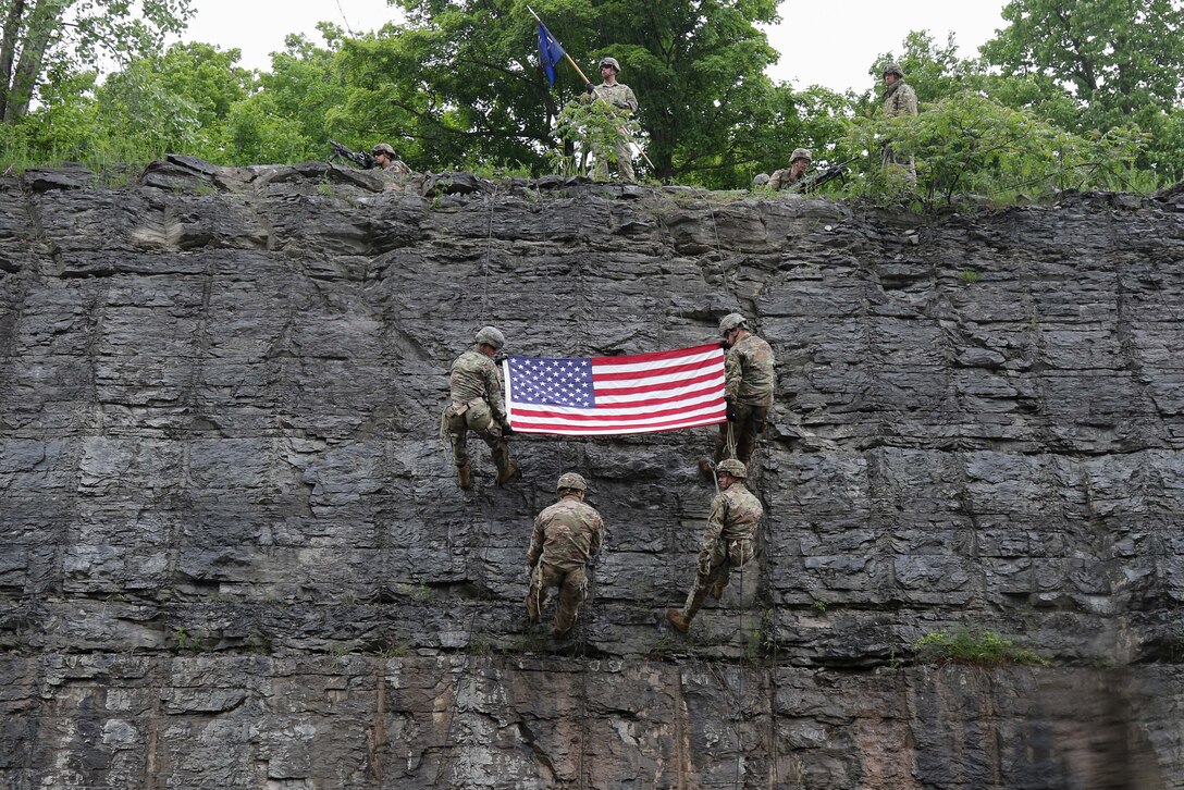 Two soldiers holding an American flag rappel down a rock face with others as more soldiers look on from the cliff top.