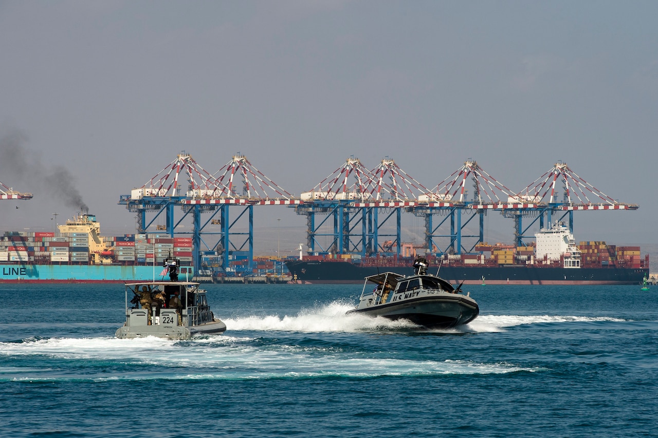 Two naval vessels speed through the water near a port. In the background are a half-dozen port cranes moving cargo from container ships.