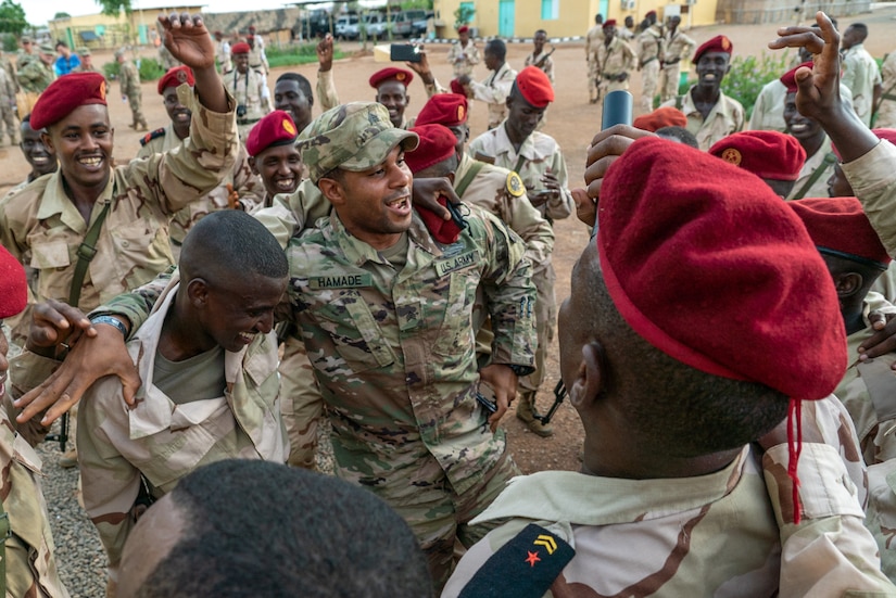 About a dozen service members stand in a loose group, outdoors, and are celebrating.