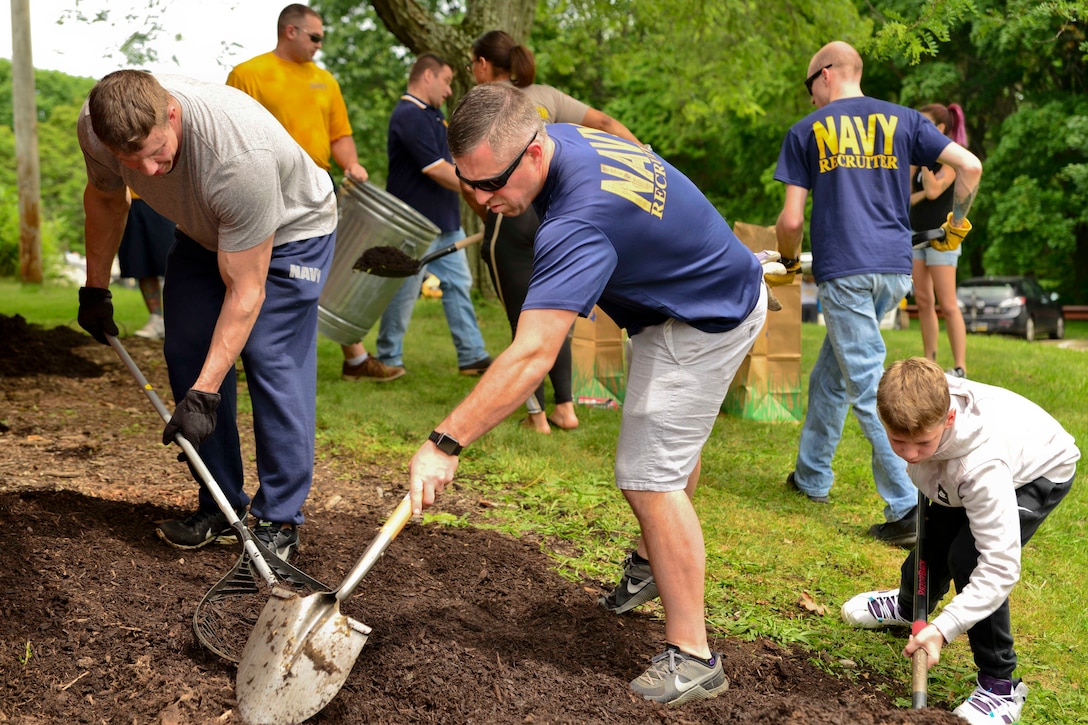 Sailors and volunteers shovel and rake mulch in a park.