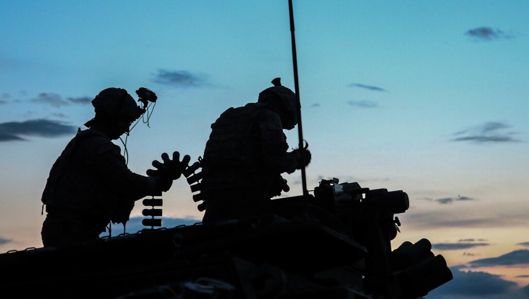 Two soldiers stand and inspect ammunition rounds against a twilight sky.