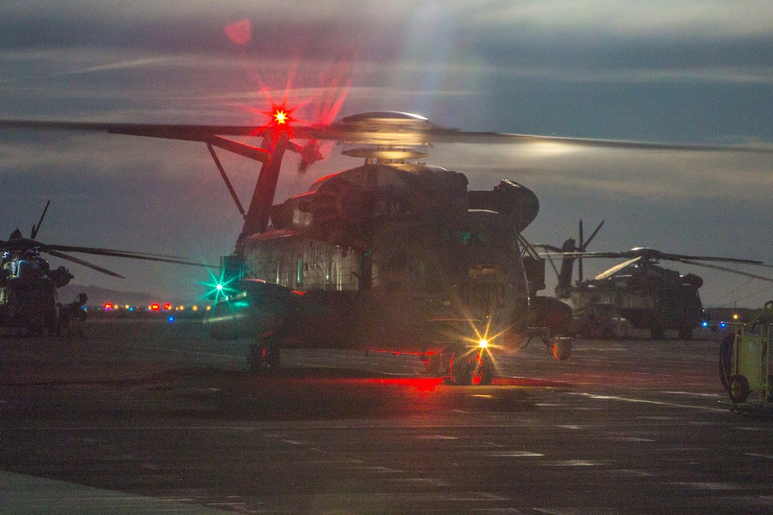 A helicopter sits on the ground with its lights on at twilight.