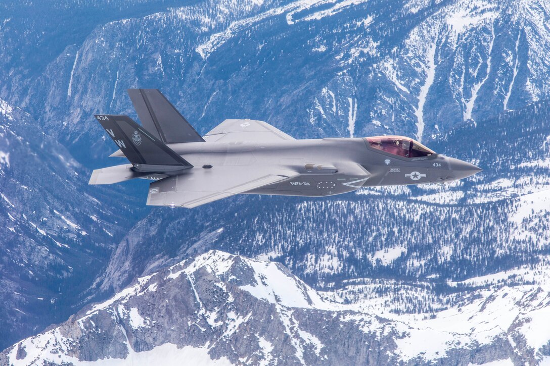 A fighter jet flies over a snowcapped mountain range.