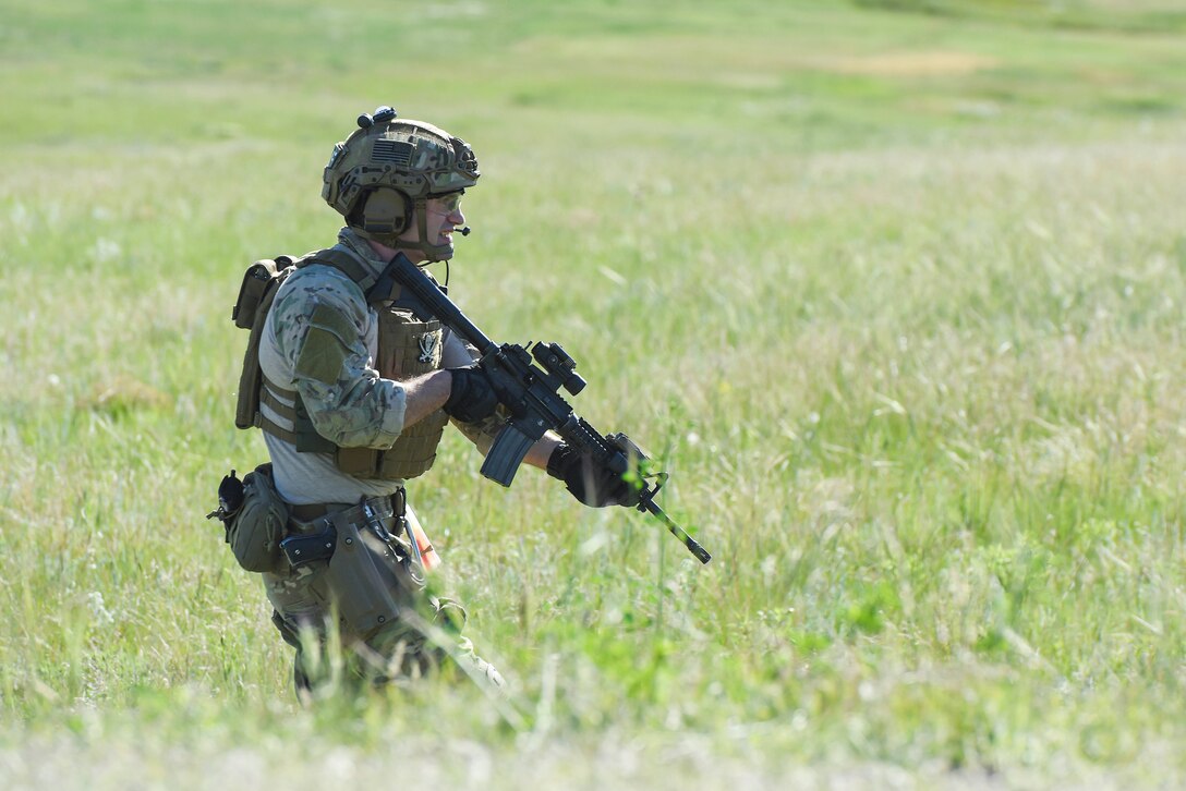 An airman wearing a helmet and carrying an automatic weapon walks through a field of tall grass.