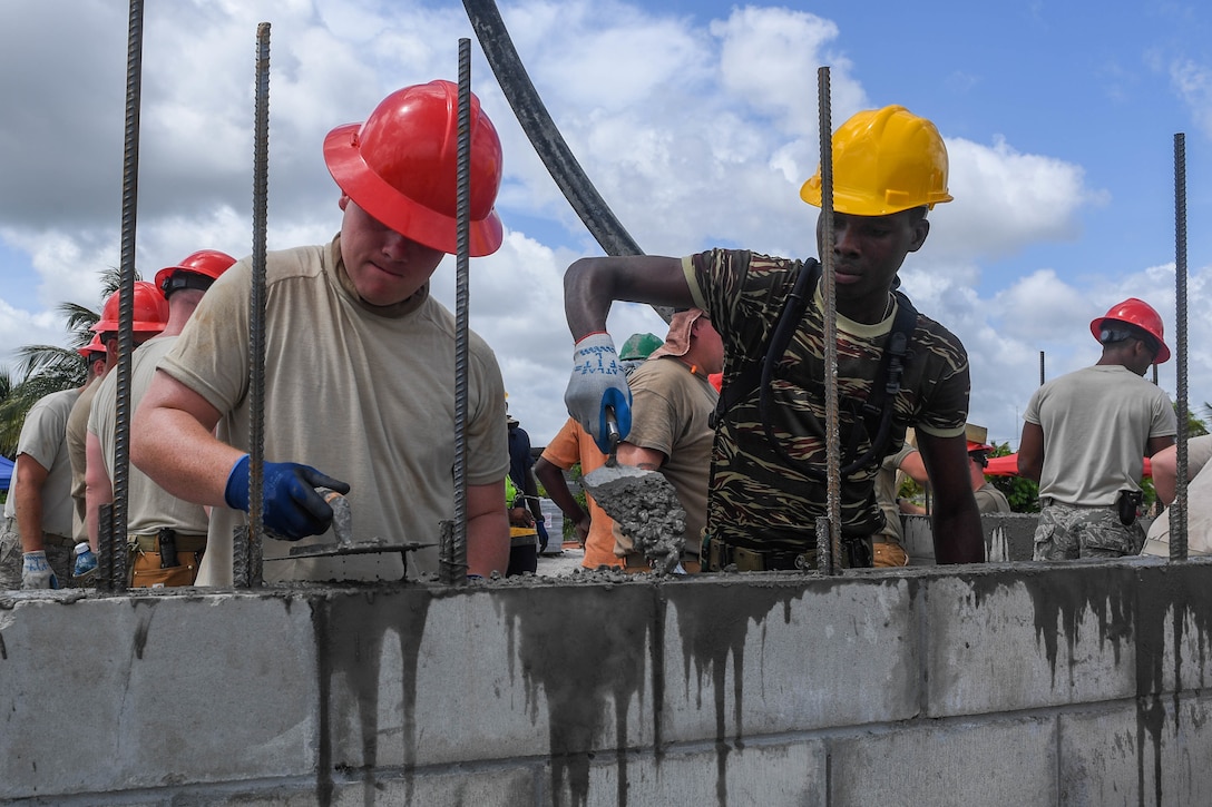 An airman and a Guyanese service member fill holes use pavers to fill holes in a concrete wall.