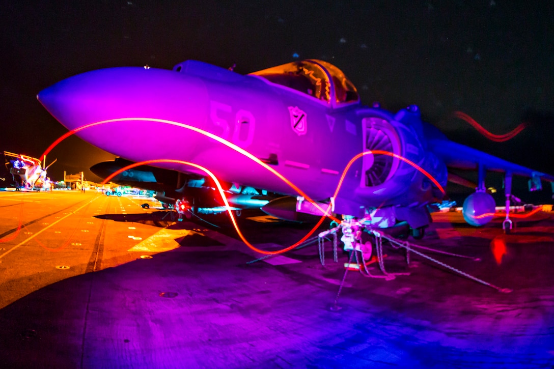 An aircraft sits on a flight deck at night as light from various machinery swirls nearby.