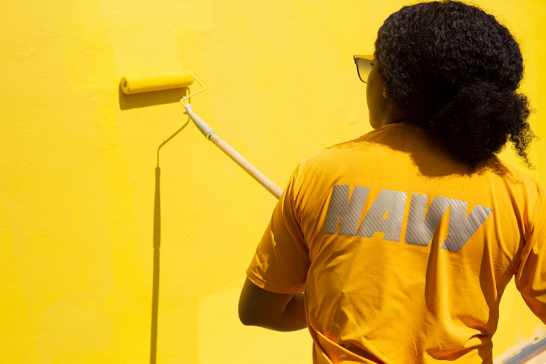 A sailors uses a roller to paint a building yellow.