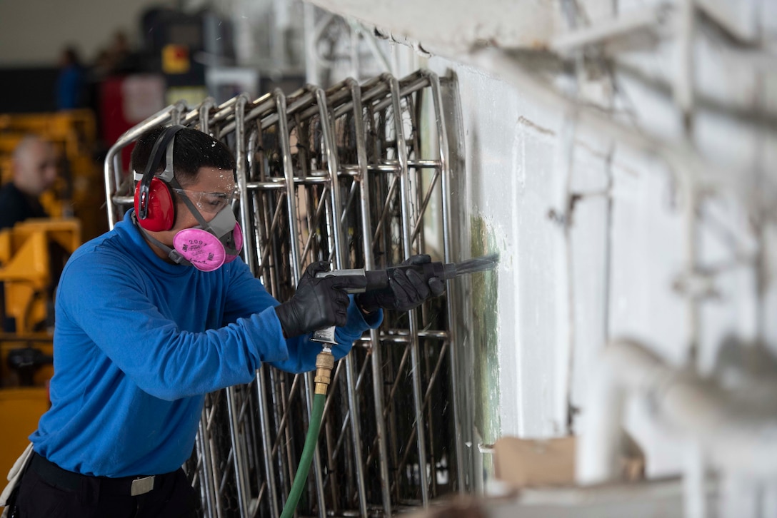 A sailor wearing safety gear uses a power tool on a partition.