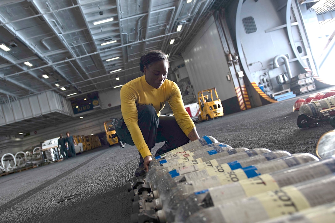 A female sailor kneels while checking a row of containers on a military ship.