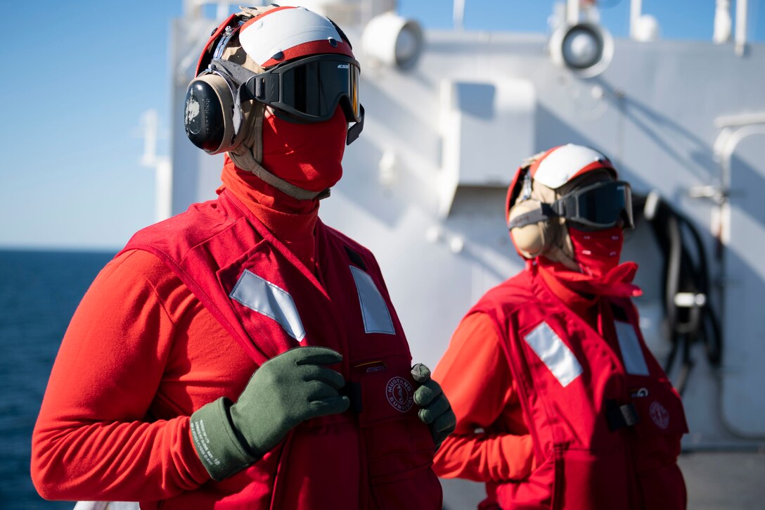 Sailors wearing goggles, earphones, red vests and scarves stand on a ship’s flight deck.