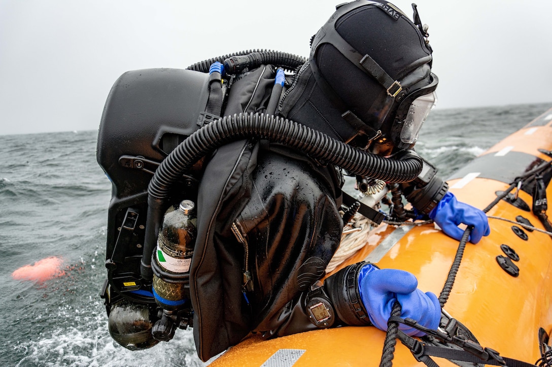 A half-submerged diver in a wetsuit carrying an oxygen tank holds on to a rope alongside a yellow raft.