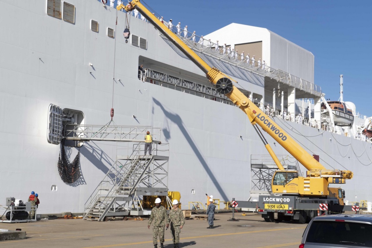 A large yellow crane rises to the deck of a hospital ship as sailors man the rails.