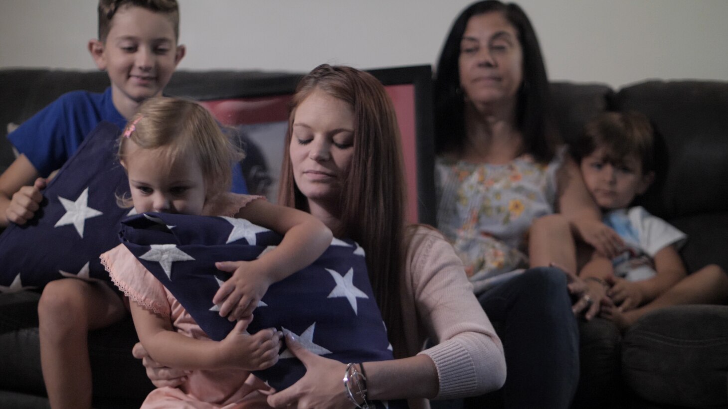 Members of the Kinkton family sits on a sofa. Morgan, wife of Kody sits on the floor with Alli in her lap who is holding a folded American flag.