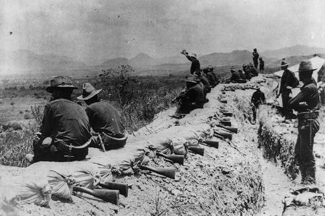 Soldiers in wide-brimmed hats sit atop trenches topped with sandbags and rifles ready for firing; one man standing in the center waves his hat.