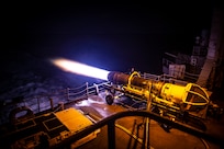 Flames spew from the back end of a jet cell being tested at night on an aircraft carrier.