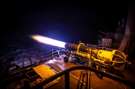 Flames spew from the back end of a jet cell being tested at night on an aircraft carrier.