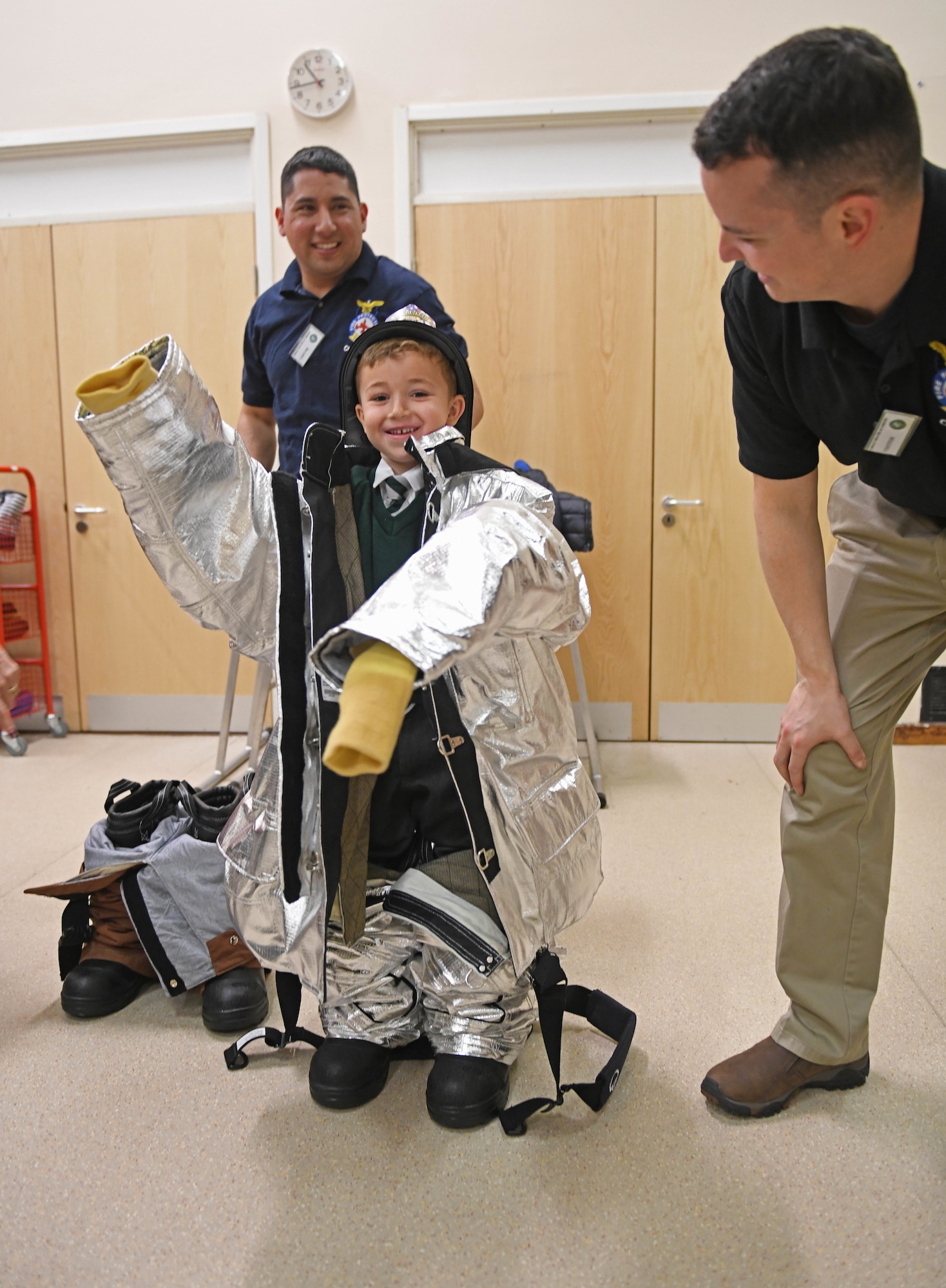 U.S. Air Force Tech. Sgt. Daniel Howard and Sergio Villela, 100th Civil Engineer Squadron firefighters, help a student put on firefighting equipment during a fire safety demonstration at Great Heath Academy, Mildenhall, England, June 19, 2019. The firefighters showed students how they use their gear and allowed them to try it on. (U.S. Air Force photo by Senior Airman Luke Milano)