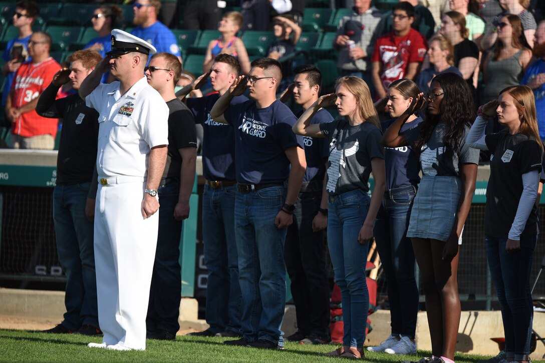 Chief Petty Officer Nathaniel Booher, of the Fargo Military Entrance Processing Station (MEPS), far left, leads a group of recent enlistees in various branches of the U.S. Military as they salute the flag during the national anthem following a commitment ceremony at the Military Appreciation Night for the Fargo-Moorhead RedHawks baseball game, Newman Outdoor Field, Fargo, N.D., June 18, 2019.