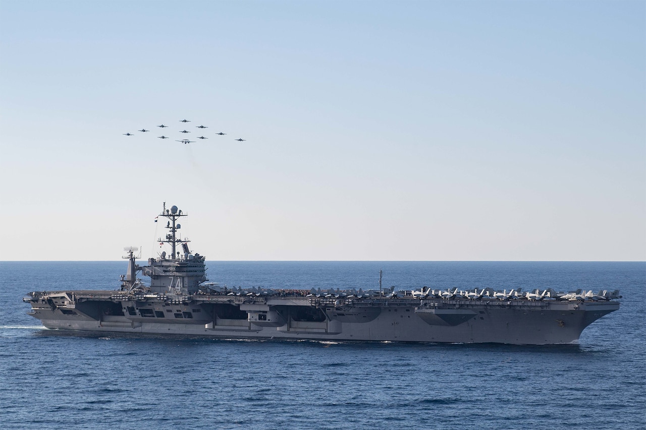 An aircraft carrier moves through the ocean. In the sky above, 11 aircraft fly in a diamond formation.