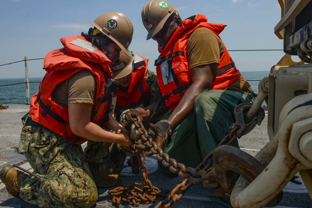 Three sailors use a metal chain to secure a vehicle to the floor.