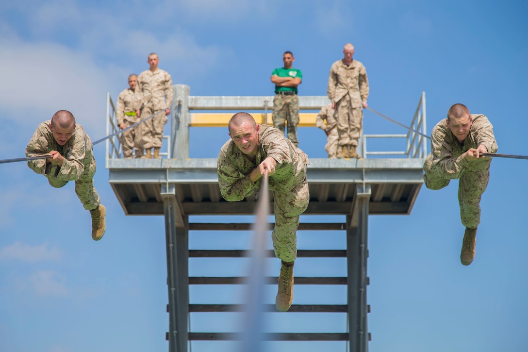 Three Marines slide along a rope suspended high above the ground as other Marines watch and wait.