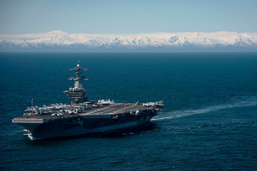 An aircraft carrier moves though the ocean. In the background are snow-covered mountains.
