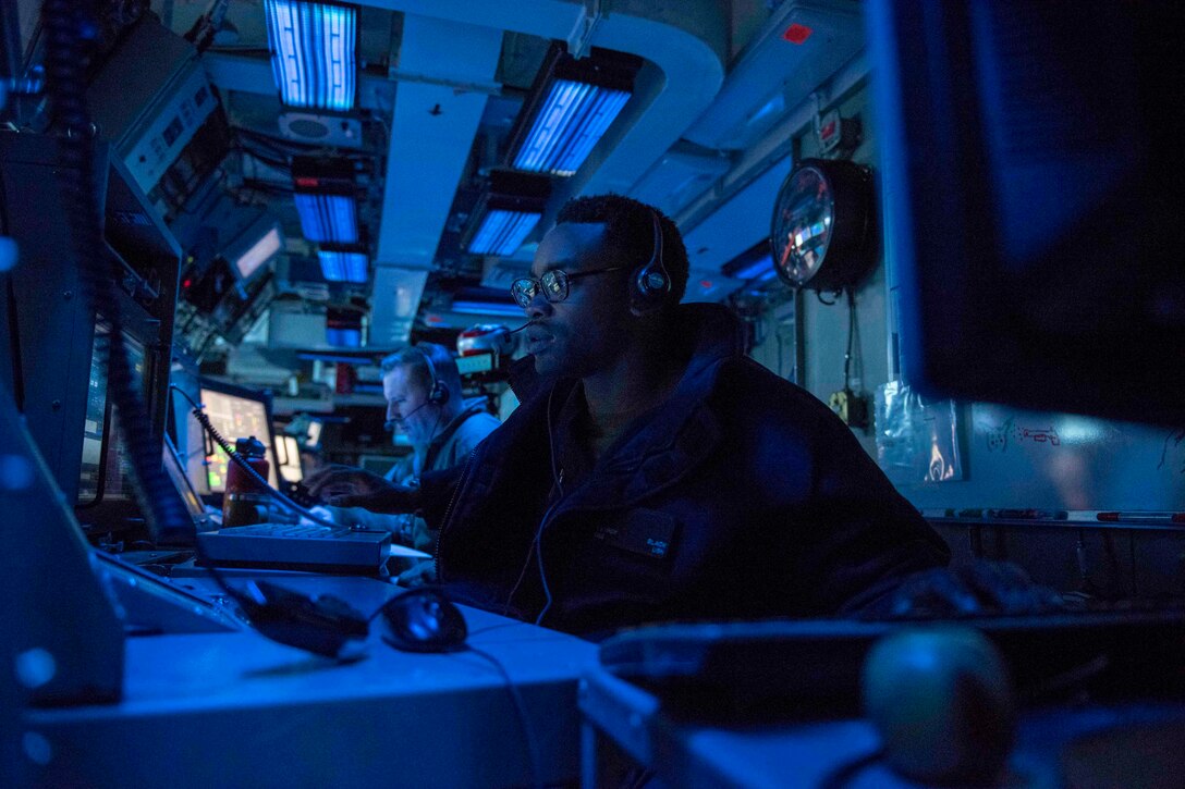 A sailor sits at a desk in front of monitors surrounded by blue light.